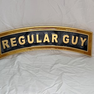 May include: A curved sign with the words "REGULAR GUY" in gold lettering. The sign has a black background with a gold glitter border. The sign is on a white background.