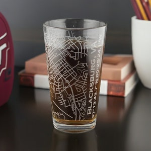 May include: A clear glass pint glass etched with a map of Blacksburg, VA, including the city's coordinates. The glass contains a dark liquid. A maroon football and a white ceramic cup with pencils are in the background.