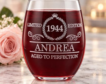May include: A stemless wine glass filled with red liquid, featuring an etched design that reads LIMITED EDITION 1944, the name ANDREA, and the phrase AGED TO PERFECTION, surrounded by a laurel wreath and a small crown icon.