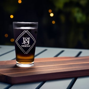 Custom laser-etched 16 oz pint glass with J&J Designz monogram on a chic outdoor patio table, perfect for personalized gifts or promotional drinkware.