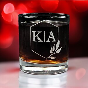 Custom laser-etched 11oz rocks glass with personalized monogrammed whiskey glass with initials 'KIA' and laurel leaf design on reflective surface with a red bokeh background, ideal for custom gift or elegant barware.