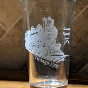 Custom-engraved 16 oz pint glass with a detailed vintage 1936 train design and initials JJK, showcasing the high-quality laser etching technique on transparent glassware.  Perfect for birthdays or special event gifts.