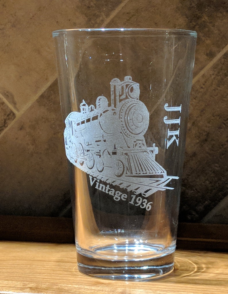 Custom-engraved 16 oz pint glass with a detailed vintage 1936 train design and initials JJK, showcasing the high-quality laser etching technique on transparent glassware.  Perfect for birthdays or special event gifts.