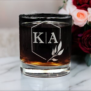 Custom laser-etched 11oz rocks glass with personalized monogrammed whiskey glass with initials 'KIA' and laurel leaf design on reflective surface with a red bokeh background, ideal for custom gift or elegant barware.