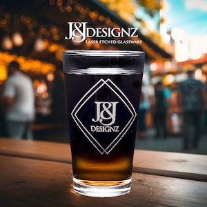 Custom-engraved 16 oz pint glass featuring J&J Designz logo with crisp laser etching, set against a lively market backdrop with warm, bokeh lighting - perfect for branded corporate gifts or personalized barware.