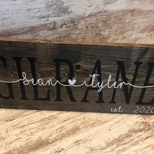 May include: A rustic wooden sign with the text "Gilrane" in black lettering. The names "Sean" and "Tyler" are written in white cursive script with a small white heart between them. The text "est 2020" is written in white below the names.