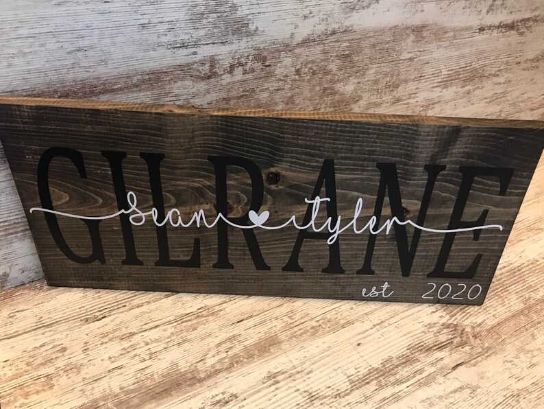 May include: A rustic wooden sign with the text "Gilrane" in black lettering. The names "Sean" and "Tyler" are written in white cursive script with a small white heart between them. The text "est 2020" is written in white below the names.