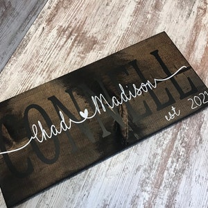 May include: A dark brown wooden sign with the text "CONNELL chad&Madison est 2022" in white lettering. The text is in a cursive font and includes a small heart symbol.