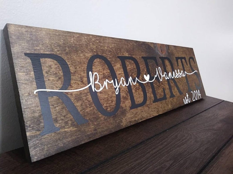 May include: A rustic wooden sign with the last name "Roberts" in black lettering. The names "Bryan" and "Vanessa" are written in white cursive with a small heart between them. The sign also includes the text "est 2014" in black lettering.