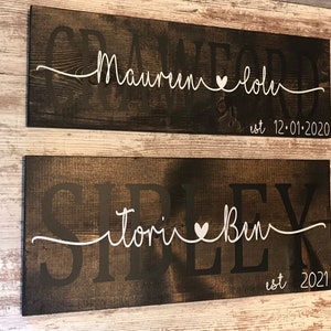 May include: Two rustic wooden signs with white lettering. The top sign reads "Crawford est 12-01-2020" with a heart between the names "Maureen" and "John". The bottom sign reads "Sirlex est 2021" with a heart between the names "Tori" and "Ben".