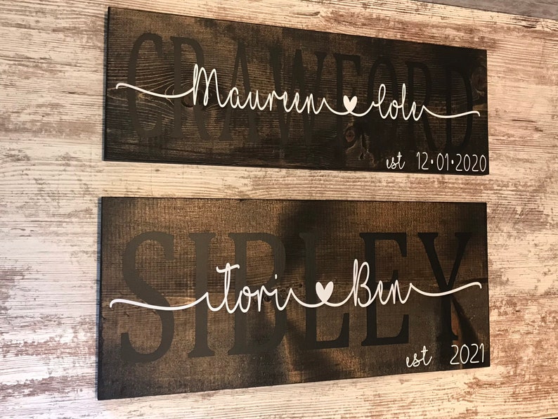 May include: Two rustic wooden signs with white lettering. The top sign reads "Crawford est 12-01-2020" with a heart between the names "Maureen" and "John". The bottom sign reads "Sirlex est 2021" with a heart between the names "Tori" and "Ben".