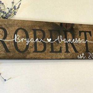 May include: A rustic wooden sign with the last name "ROBERTS" in large black letters. The names "Bryan" and "Vanessa" are written in cursive with a small white heart between them. The text "est 2014" is written in white below the names.