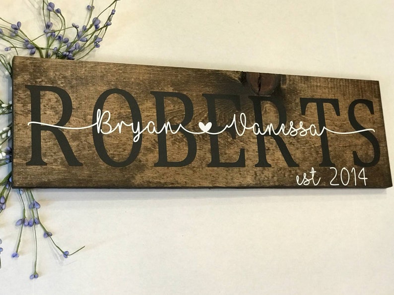 May include: A rustic wooden sign with the last name "ROBERTS" in large black letters. The names "Bryan" and "Vanessa" are written in cursive with a small white heart between them. The text "est 2014" is written in white below the names.