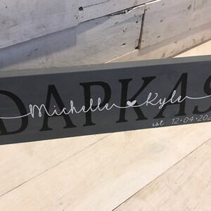 May include: Gray wooden sign with the text "Darkas" in black lettering. The text "Michelle & Kyle" is written in white cursive lettering with a small heart symbol between the names. The date "12.04.2021" is written in white lettering below the names.