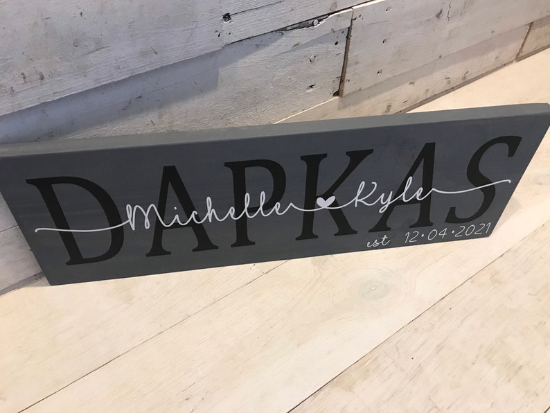 May include: Gray wooden sign with the text "Darkas" in black lettering. The text "Michelle & Kyle" is written in white cursive lettering with a small heart symbol between the names. The date "12.04.2021" is written in white lettering below the names.