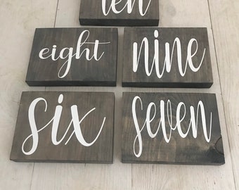 May include: Six wooden signs with white painted numbers on a light wood background. The numbers are six, seven, eight, nine, ten.