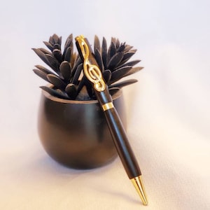 Puede incluir: Un bolígrafo negro y dorado con un diseño de clave de sol. El bolígrafo está apoyado en una maceta negra con una planta suculenta.