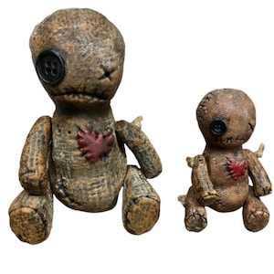 May include: Two handmade voodoo dolls. The larger doll has a button eye and a stitched mouth, with a red heart patch on its chest. The smaller doll has similar features. Both dolls are brown and tan with a distressed appearance.