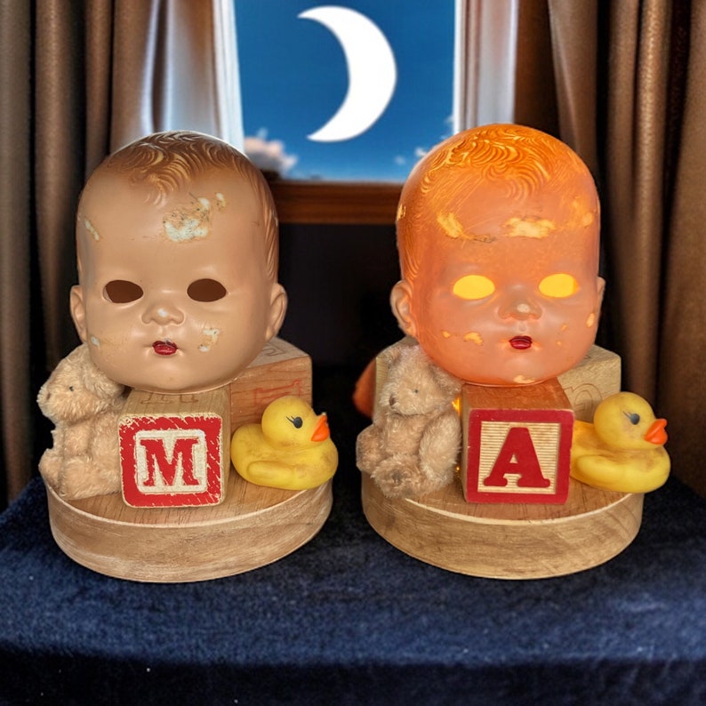May include: Two vintage baby doll head nightlights with a yellow rubber ducky and a teddy bear on each side. The nightlights are on wooden bases with a red block that says "M" on the left and "A" on the right.