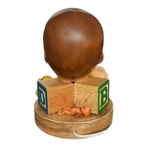 May include: A wooden lamp base with a ceramic baby head sculpture. The base is decorated with wooden blocks that spell "D", "O", "Z", and "B". There is a small baby doll lying on the base.