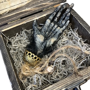 Könnte beinhalten: Eine schwarze Hand mit einem goldenen Armband befindet sich in einer Holzkiste, die mit grauem und braunem zerkleinertem Papier gefüllt ist.