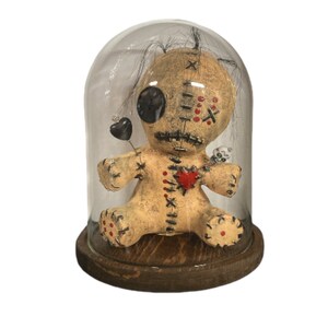 May include: A voodoo doll with a stitched heart, red and black accents, and a black button eye, is displayed under a glass dome on a wooden base.