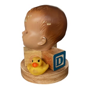 May include: A vintage doll head with a brown complexion and a yellow rubber ducky sitting on a wooden base with colorful alphabet blocks. The blocks spell out the letter "D".