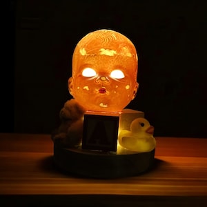 May include: A nightlight shaped like a baby doll's head with glowing eyes. The doll is sitting on a base with a toy block and a rubber duck.