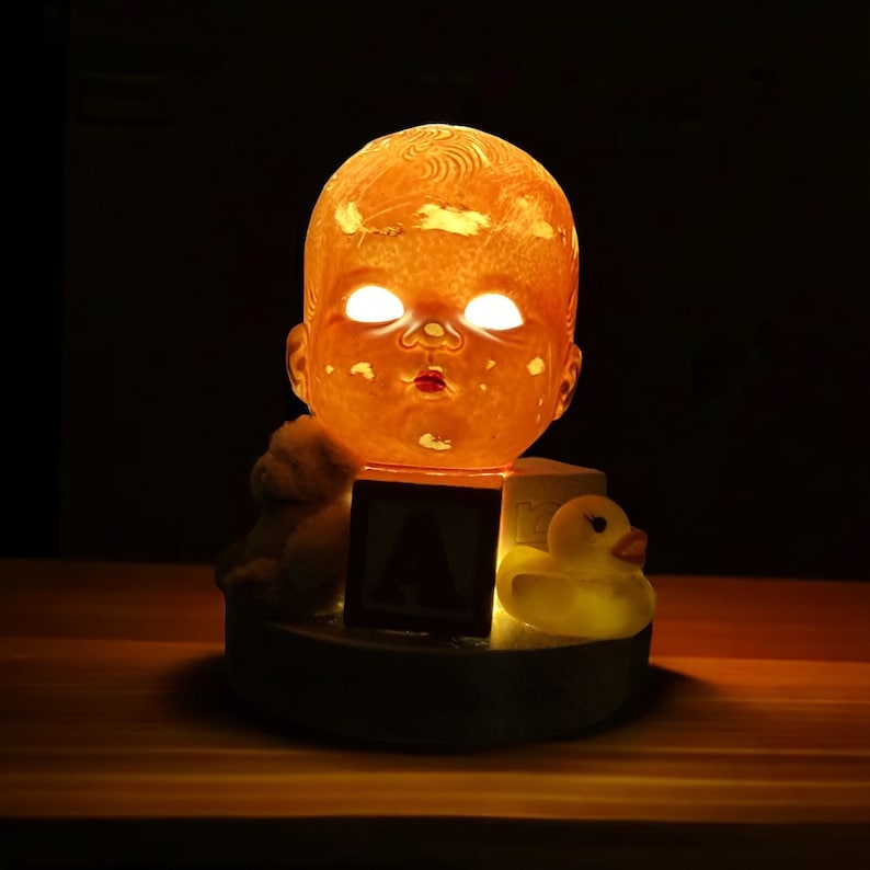 May include: A nightlight shaped like a baby doll's head with glowing eyes. The doll is sitting on a base with a toy block and a rubber duck.