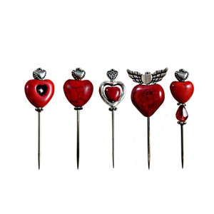May include: Set of five red heart-shaped pins with silver accents. The pins have different designs, including a heart with wings, a heart with a smaller heart inside, and a heart with a red gemstone.