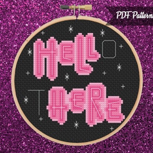 May include: A black and white cross stitch pattern with pink lettering that reads "Hello There" on a black background. The pattern is in a round embroidery hoop.