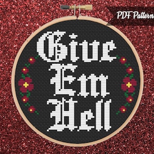 May include: A black and white cross-stitch pattern with the text "Give Em Hell" in a gothic font. The pattern is surrounded by a wreath of red flowers.