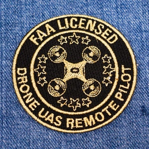 Puede incluir: Un parche bordado en negro y dorado con el texto "FAA Licensed Drone UAS Remote Pilot" y un gráfico de dron estilizado en el centro.