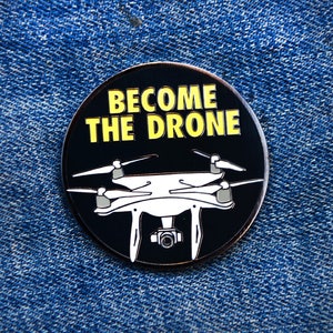 May include: A black and white enamel pin with a drone silhouette and the text "Become the Drone" in yellow letters.