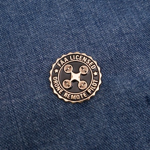 May include: Gold-colored pin with a black background. The pin features a drone icon and the text "FAA LICENSED DRONE REMOTE PILOT".