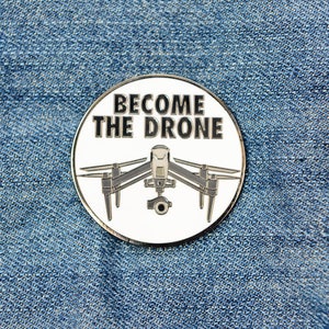 May include: A white enamel pin with a black outline. The pin features a drone with the text "Become the Drone" in black.