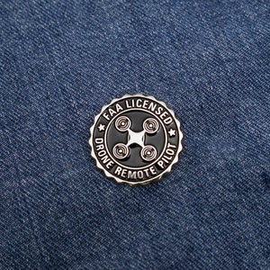 May include: A silver pin with a black background and white text. The pin says "FAA Licensed Drone Remote Pilot" and features a stylized drone icon.