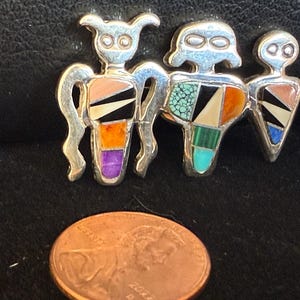 May include: A silver-toned brooch featuring three stylized figures with inlaid colorful stones. The figures have rounded heads and geometric bodies with orange, turquoise, black, and other colored stone inlays. A penny is in the foreground.