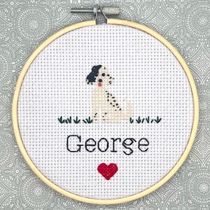 May include: A circular wooden embroidery hoop containing white Aida fabric with a cross-stitch design of a white and black spotted dog, the name George in black lettering, and a small red heart, all positioned above a row of green grass.