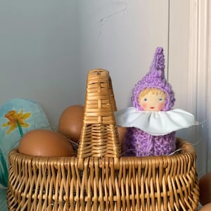 Puede incluir: Una cesta de mimbre tejida llena de huevos marrones y una muñeca hecha a mano. La muñeca lleva un traje de punto morado con cuello blanco y gorro puntiagudo. Un huevo de Pascua con un diseño de narciso también está en la cesta.
