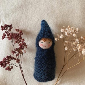 May include: A handmade doll with a blue knitted hooded cloak. The doll has a painted face with rosy cheeks and brown hair. Dried burgundy and white flowers are arranged on either side of the doll.