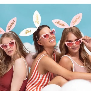 May include: Three friends wearing pink bunny ear headbands with the words "Bride" and "Babes" on them. They are also wearing pink heart-shaped sunglasses. The headband on the middle friend says "The Bride".