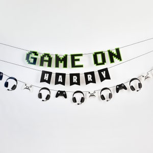 Peut inclure: Une bannière noire et verte avec les mots "GAME ON" et "HARRY" en lettres pixelisées. La bannière est décorée de manettes de jeu et de casques noirs et blancs.