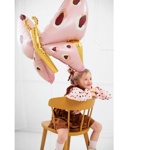 May include: A young girl sits on a yellow chair holding a pink and gold butterfly balloon. The balloon has a red and white polka dot pattern.