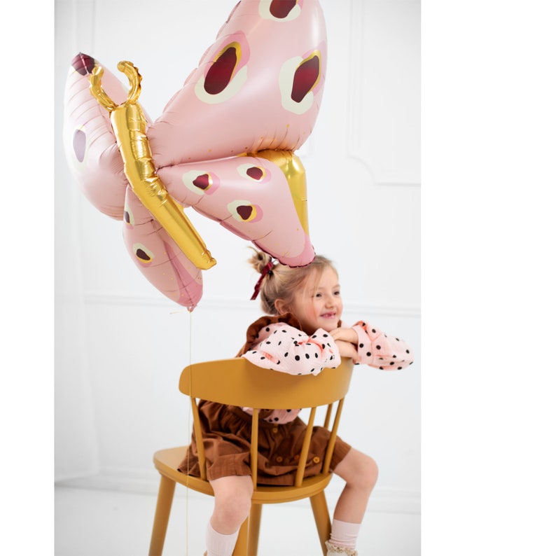 May include: A young girl sits on a yellow chair holding a pink and gold butterfly balloon. The balloon has a red and white polka dot pattern.