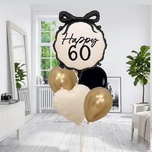 May include: A celebratory balloon arrangement for a 60th birthday. It features a large cream balloon with "Happy 60" in black script, topped with a black bow. The bouquet includes gold, black, and a cream heart-shaped balloon.