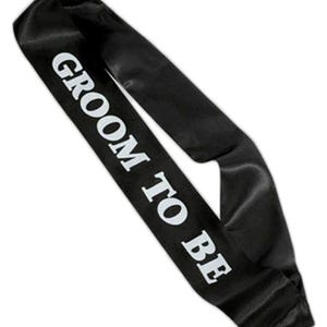 Könnte beinhalten: Schwarze Satinschärpe mit dem weißen Schriftzug "GROOM TO BE". Die Schärpe wird diagonal über den Körper getragen, ein übliches Accessoire für Junggesellenabschiede und Hochzeitsfeiern.