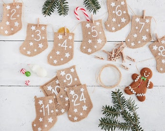 May include: A burlap advent calendar with numbers 1 through 24, decorated with white stars. The calendar is made of burlap and has a rustic look. The numbers are white and are printed on the burlap. The calendar is hanging on a white wall. There is a gingerbread man cookie and a sprig of greenery in the image.