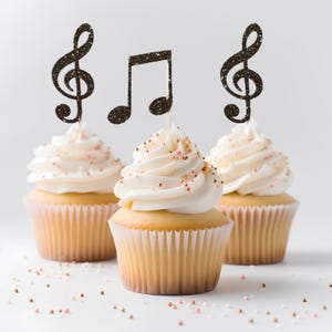 Puede incluir: Tres cupcakes con glaseado blanco, decorados con chispas doradas y rosas. Cada cupcake tiene un adorno de purpurina de una nota musical o una clave de sol. Los cupcakes están sobre una superficie blanca, con chispas esparcidas.
