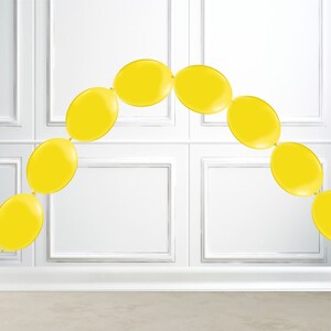 May include: A garland of bright yellow balloons arranged in an arch shape. The balloons are oval and connected, creating a festive decoration. The background is a white wall with rectangular paneling.